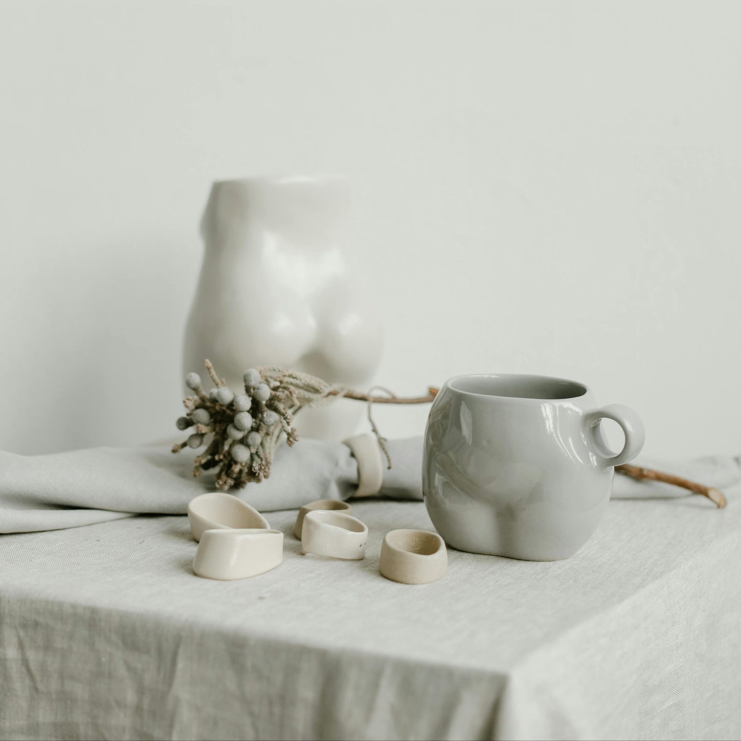 A grey mug evocative of the female form on a table setting with irregular rings, flowers, and artistic nude sculptures.