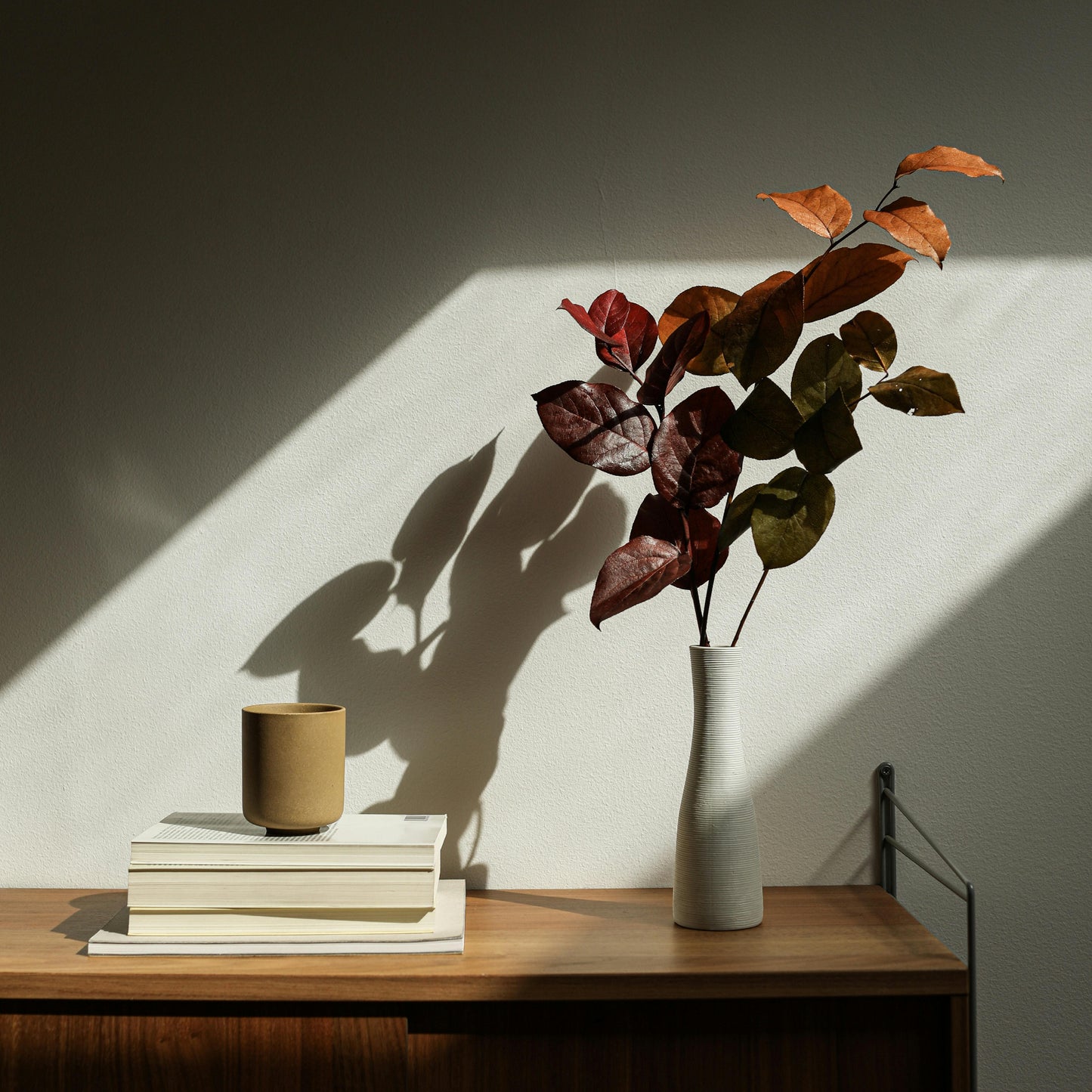 A tall, ridged white vase with a long tapered neck with a bouquet of red leaves in bright sunlight.
